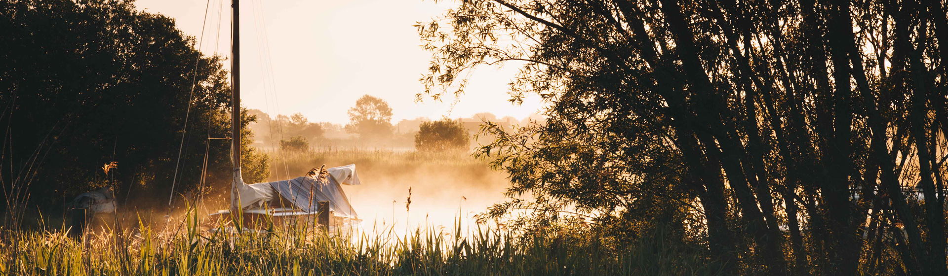 Early morning sunlight casts a warm golden glow over Upton Dyke, where a small boat covered with a tarp rests beside tall reeds. Mist rises gently from the still water, and trees frame the scene in soft silhouette, creating a peaceful, atmospheric riverside view.
