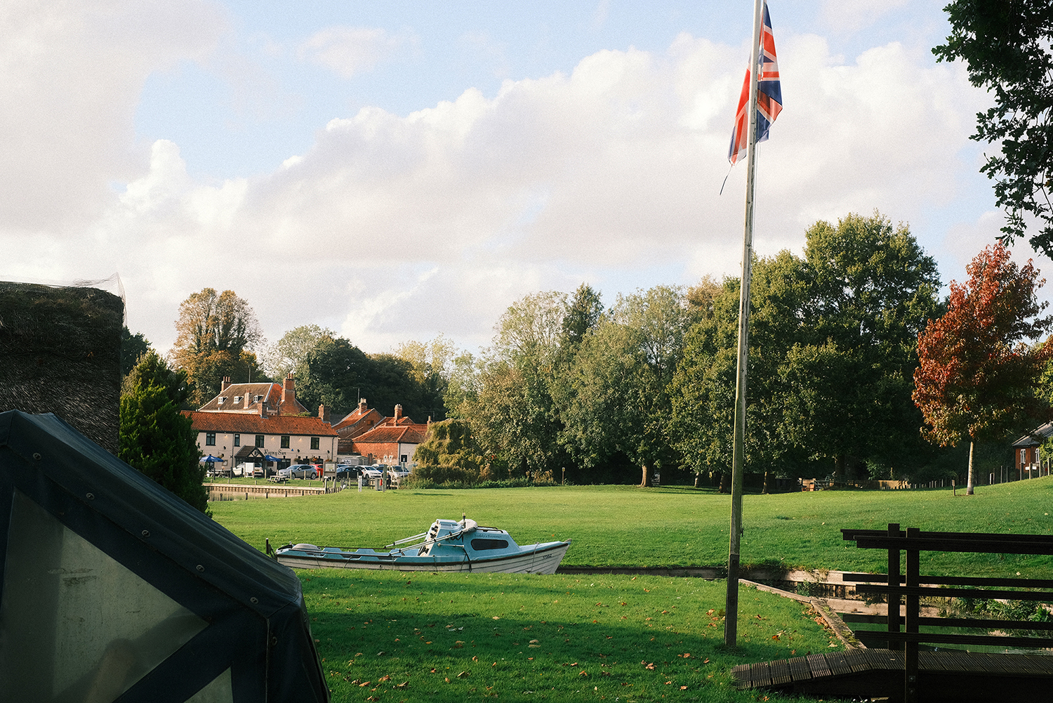 A typical Broads scene at Coltishall Common, by Tom Waterfall A grassy riverside common with a small blue-and-white boat moored along the water’s edge, a Union Jack flag flying on a tall pole, and a backdrop of trees in early autumn colors. In the distance, traditional red-roofed buildings and parked cars can be seen under a partly cloudy sky. Ask Cha