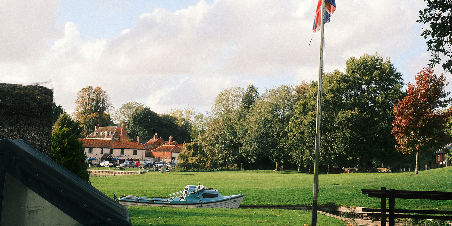 A grassy riverside common with a small blue-and-white boat moored along the water&rsquo;s edge, a Union Jack flag flying on a tall pole, and a backdrop of trees in early autumn colors. In the distance, traditional red-roofed buildings and parked cars can be seen under a partly cloudy sky.          Ask Cha