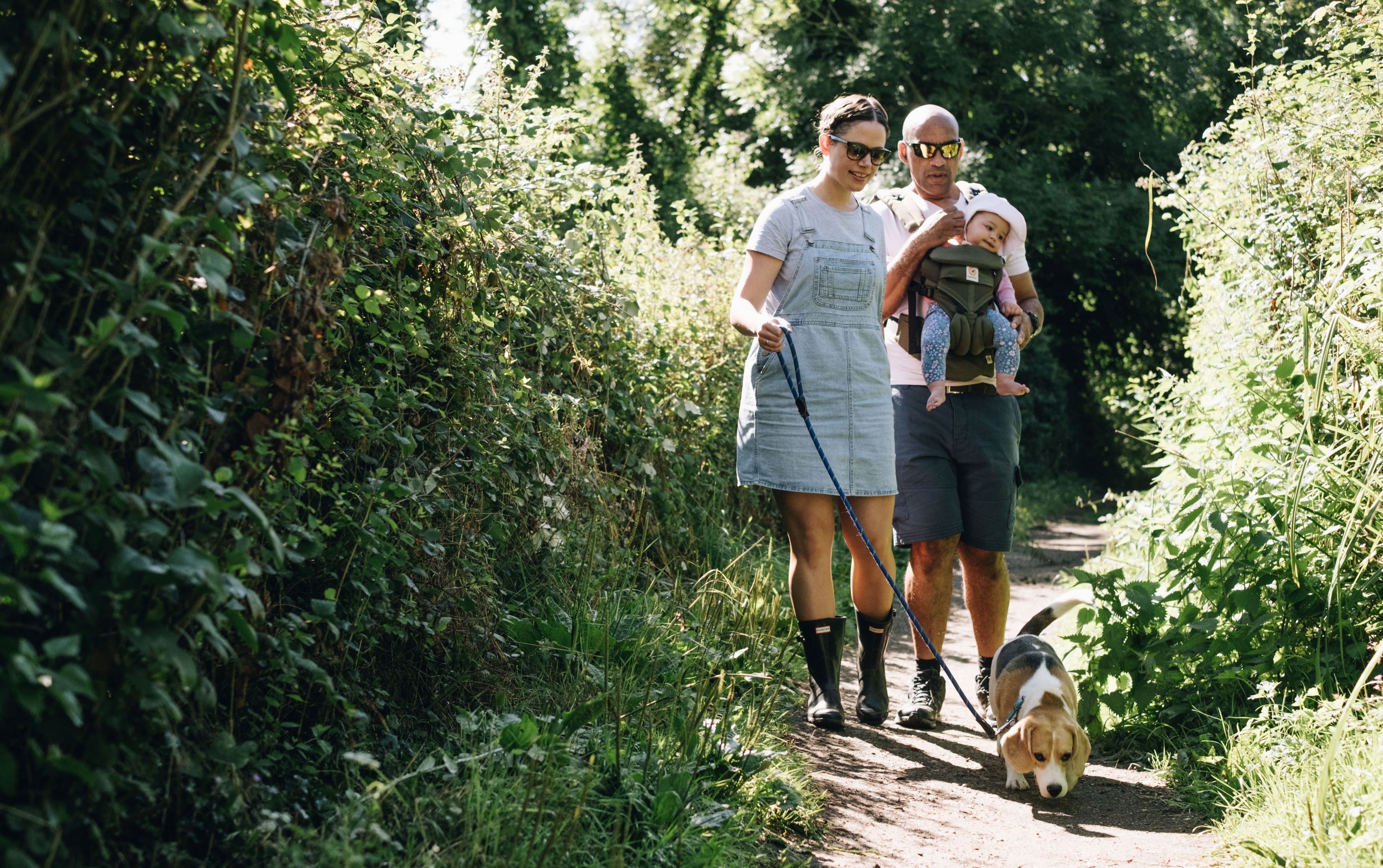 Two adults walking along a narrow, leafy path with a dog on a lead; one adult is carrying a baby in a front carrier. Sunlight filters through the trees.
