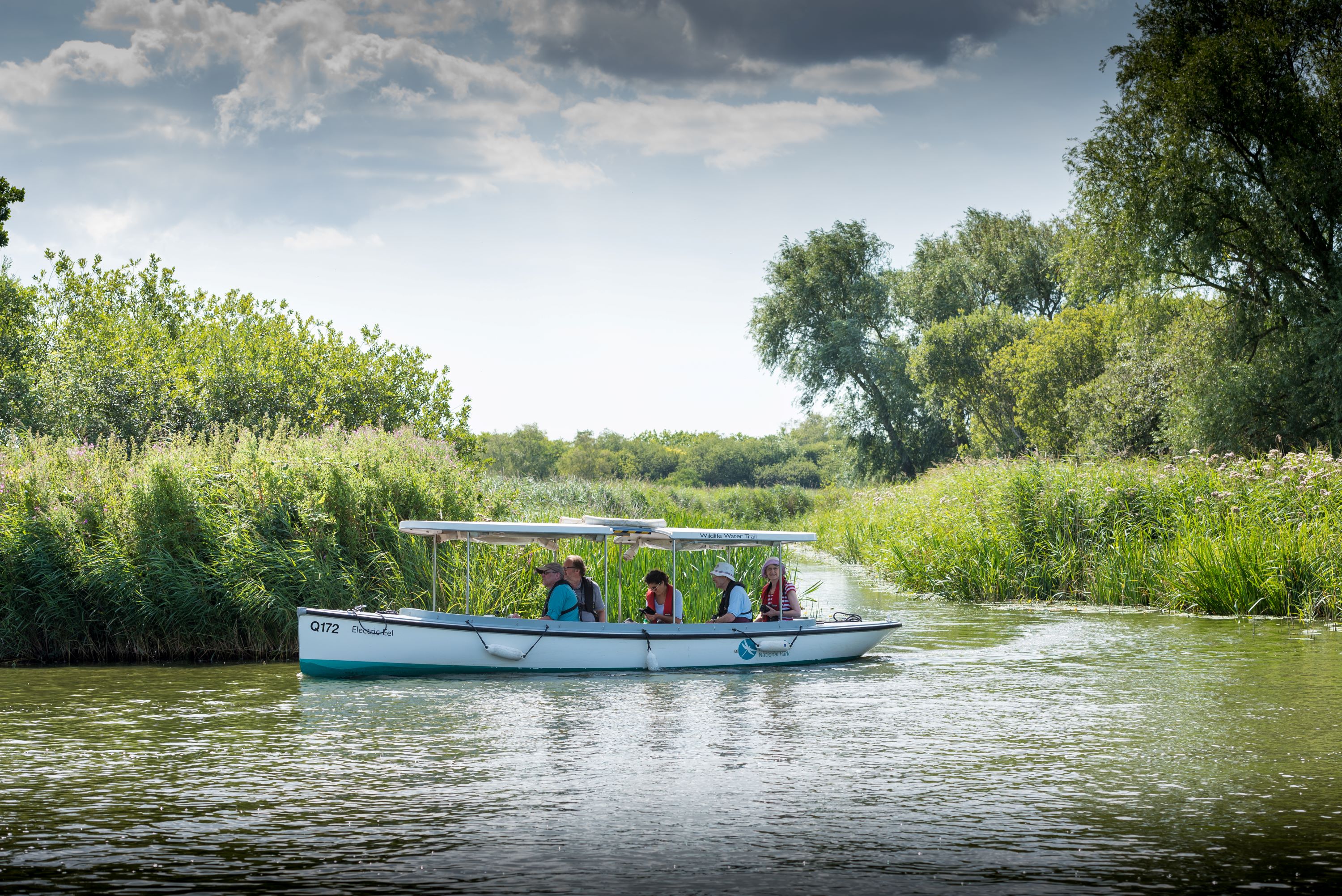A small passenger boat carrying several people travels along a calm river, surrounded by tall green reeds and trees under a partly cloudy sky.