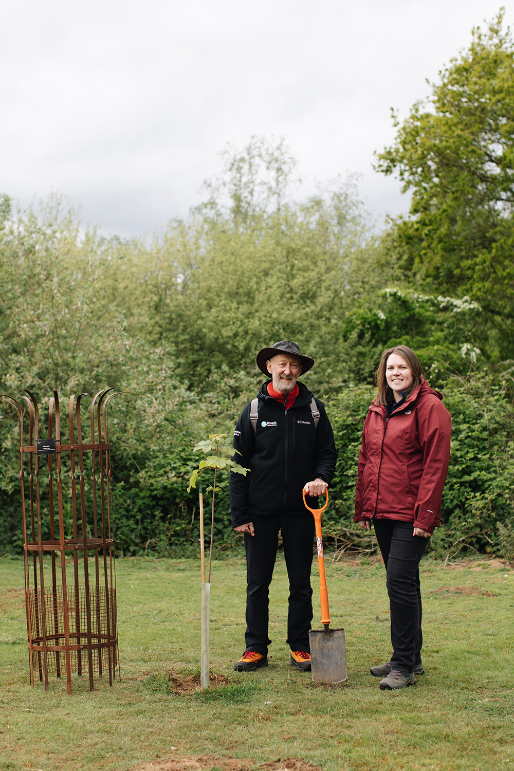 John Packman and Lynsey Skeates alongside the planted sapling Two people stand in a green outdoor setting beside a newly planted young tree and a metal tree guard; one holds a spade, with dense trees and bushes behind them.