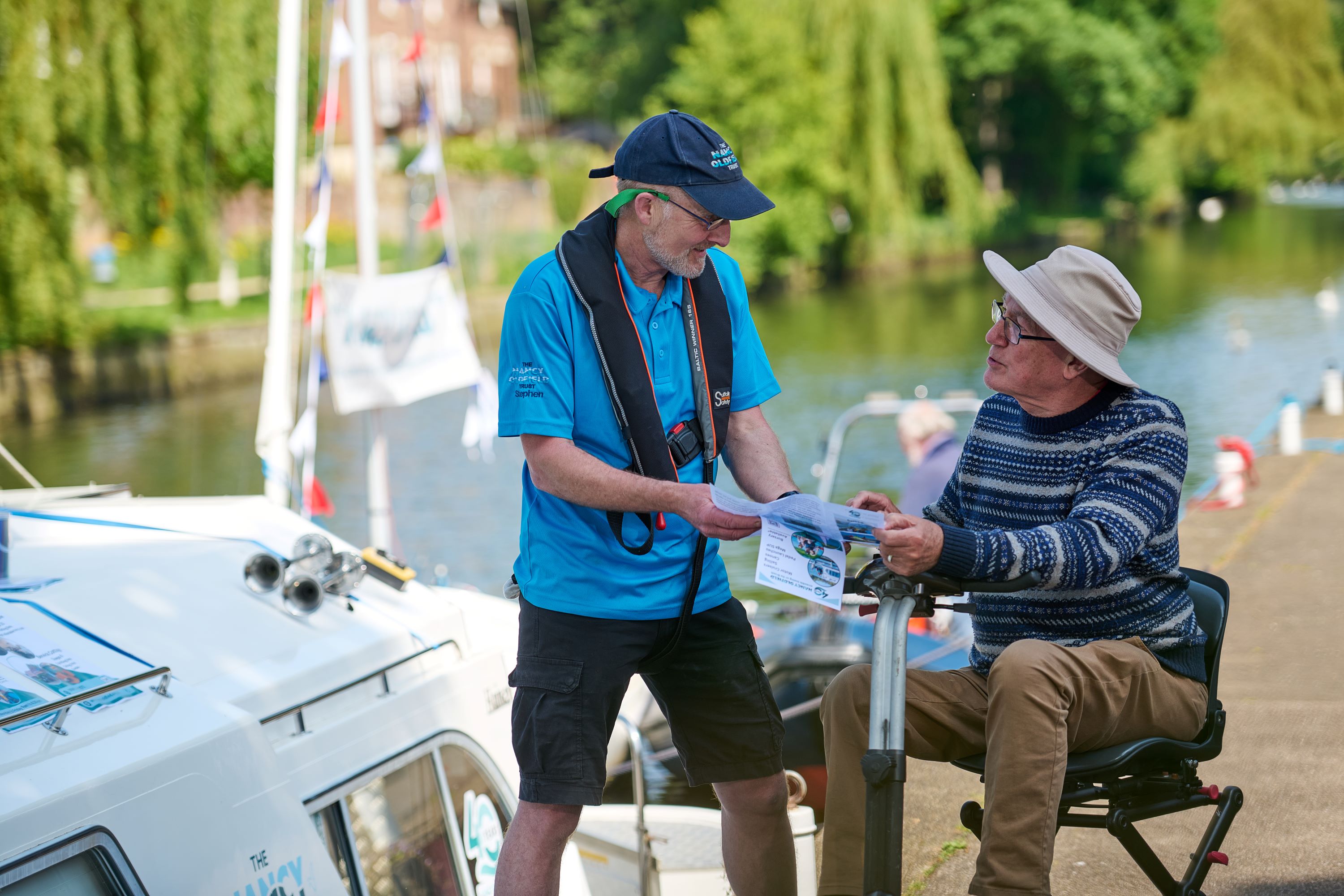 A Quay Ranger in a blue polo shirt and life jacket hands a leaflet to an older person seated on a mobility scooter beside a canal. A small boat is moored nearby, with bunting and greenery along the waterside in the background.