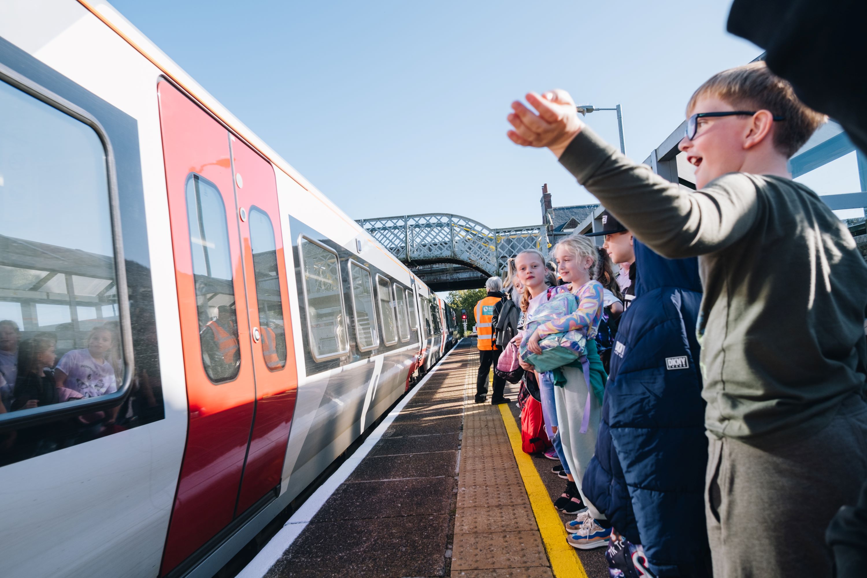 Oulton Broad Primary School students catching the train People stand along a station platform as a red-and-white train pulls in, with a railway bridge visible ahead. A staff member in a high‑visibility vest stands near the doors while passengers wait behind the yellow line on a sunny day.