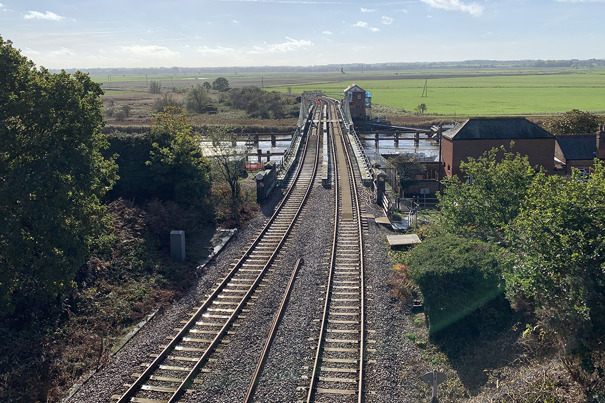 Railway tracks stretch across a bridge over a river in a flat rural landscape. The tracks lead towards open green fields on the horizon, passing a small signal or control building beside the bridge. Trees and vegetation line both sides of the railway in the foreground, while sunlight reflects on the water below. The scene is viewed from an elevated position under a bright blue sky with light cloud.