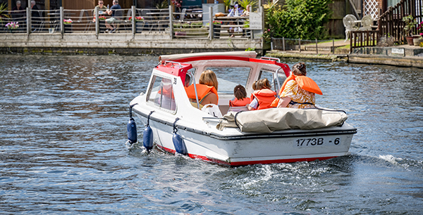 A small white and red day boat carrying four people, all wearing bright orange life jackets, cruises along a river. Two adults and two children sit at the back, enjoying the ride. In the background, a riverside caf&eacute; with people sitting at tables and a wooden deck is visible