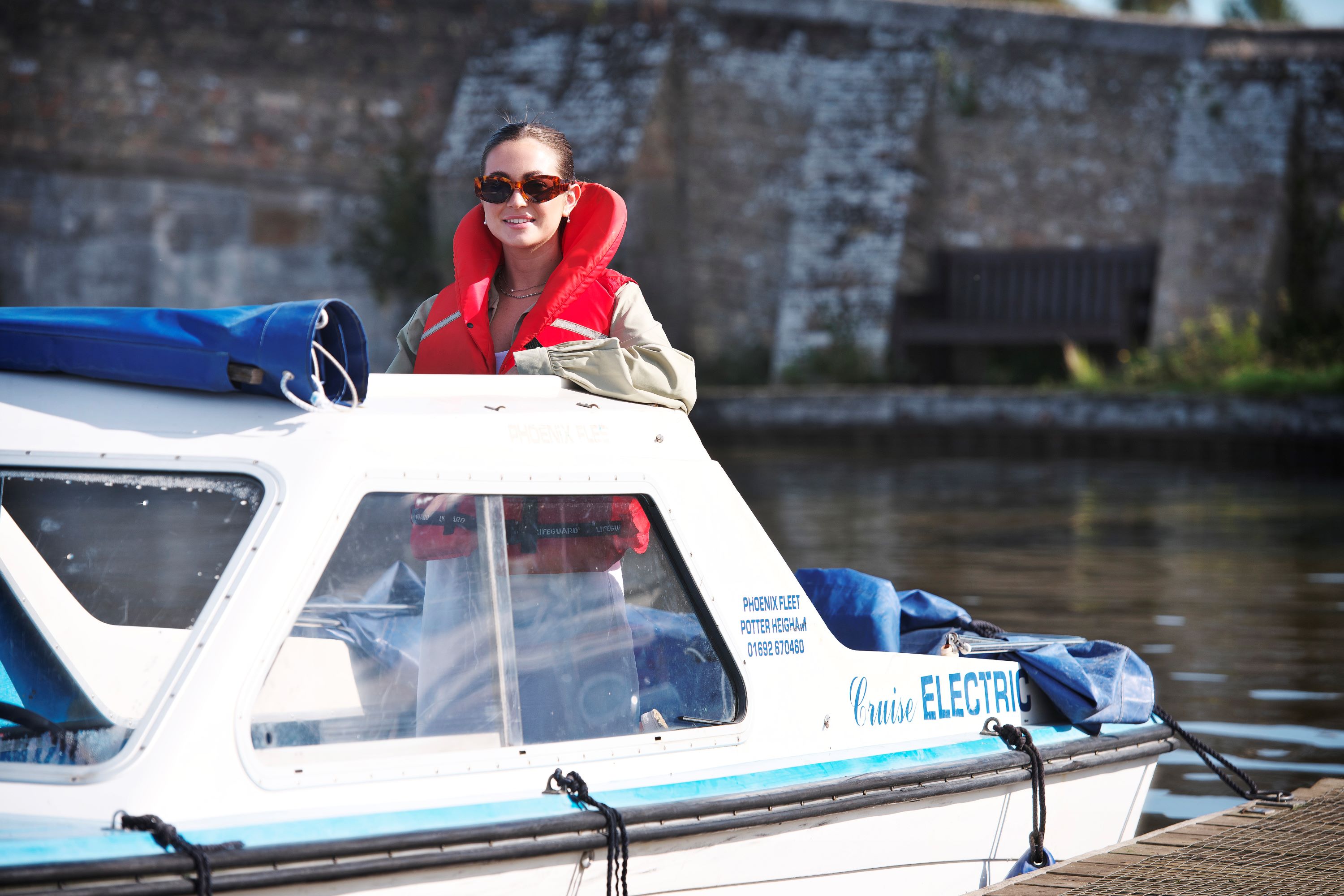Day boats at Potter Heigham A person wearing a red life jacket stands on a small white electric boat moored on a calm river. The boat is tied to the bank, with a stone wall and water in the background on a bright day.
