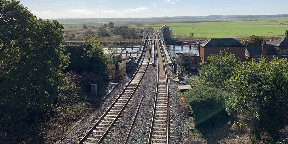 Railway tracks stretch across a bridge over a river in a flat rural landscape. The tracks lead towards open green fields on the horizon, passing a small signal or control building beside the bridge. Trees and vegetation line both sides of the railway in the foreground, while sunlight reflects on the water below. The scene is viewed from an elevated position under a bright blue sky with light cloud.