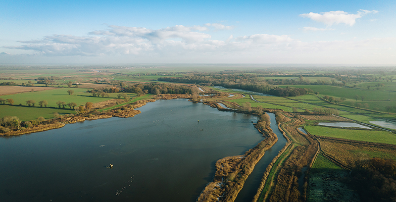 An image shows an aerial view of the Broads&rsquo; wetland landscape. A large body of water is surrounded by patches of green fields, marshes, and winding water channels. The horizon stretches far, under a partly cloudy sky, giving the scene a peaceful and expansive feel.