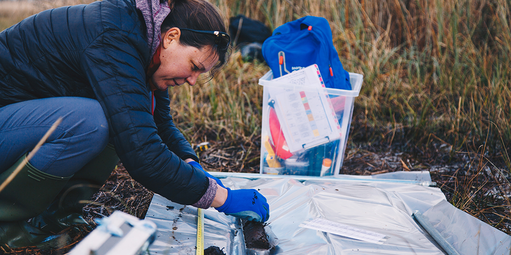 A person wearing outdoor clothing and blue gloves kneels on grassland, measuring and examining a soil sample laid out on a plastic sheet, with field equipment and a backpack nearby.
