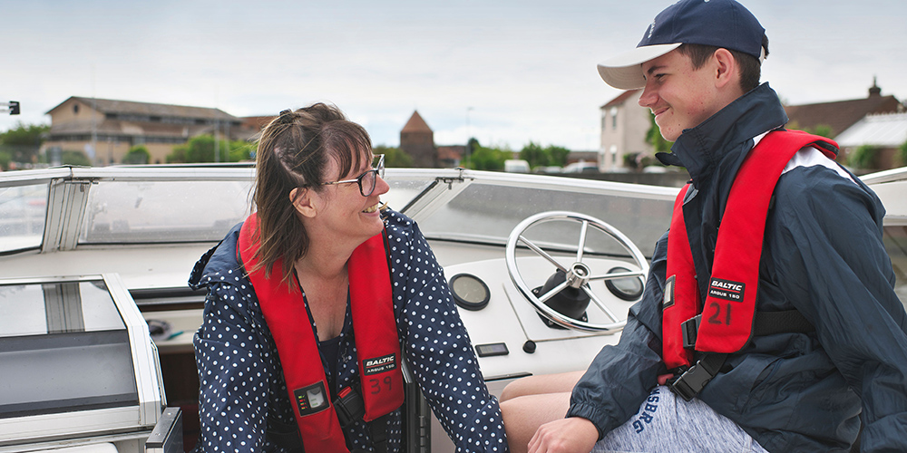 d4dbdea6f9eae532ad91afb49aab889604184f03.jpg Two people wearing red life jackets sit aboard a boat and talk near the helm, with riverside buildings visible in the background.