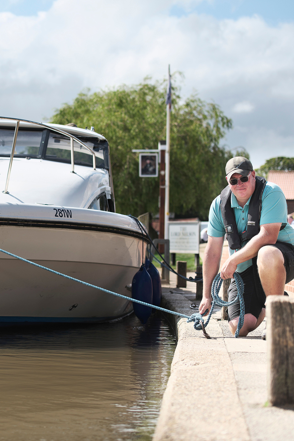 A Broads Authority Quay Ranger at Reedham Person kneeling on a quay secures a rope from a moored motorboat, wearing a life jacket and cap beside calm water.