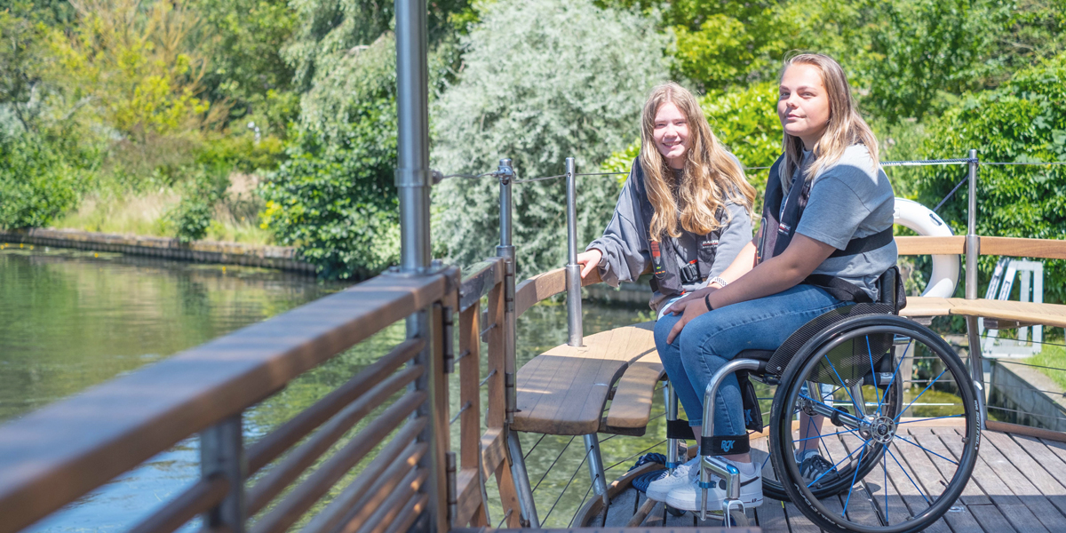 Two young visitors in wheelchairs on a wooden boardwalk looking over Barton Broad 