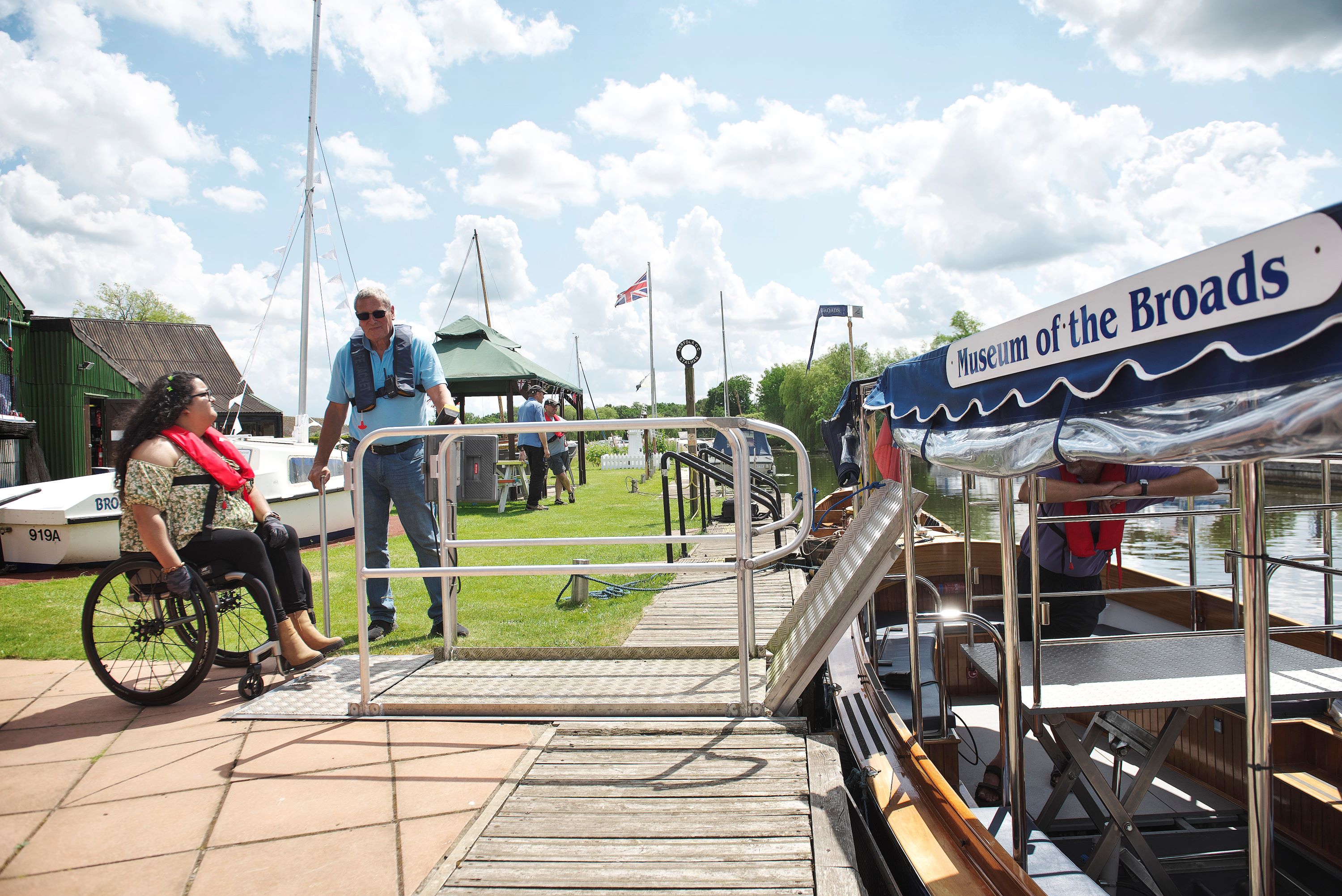 Boarding the accessible trip boat at the Museum of the Broads A wheelchair user waits on a paved riverside area while a staff member stands by a metal boarding ramp leading onto a moored boat. Other boats, grassy banks, and a canopy marked “Museum of the Broads” are visible along the water on a bright, partly cloudy day.
