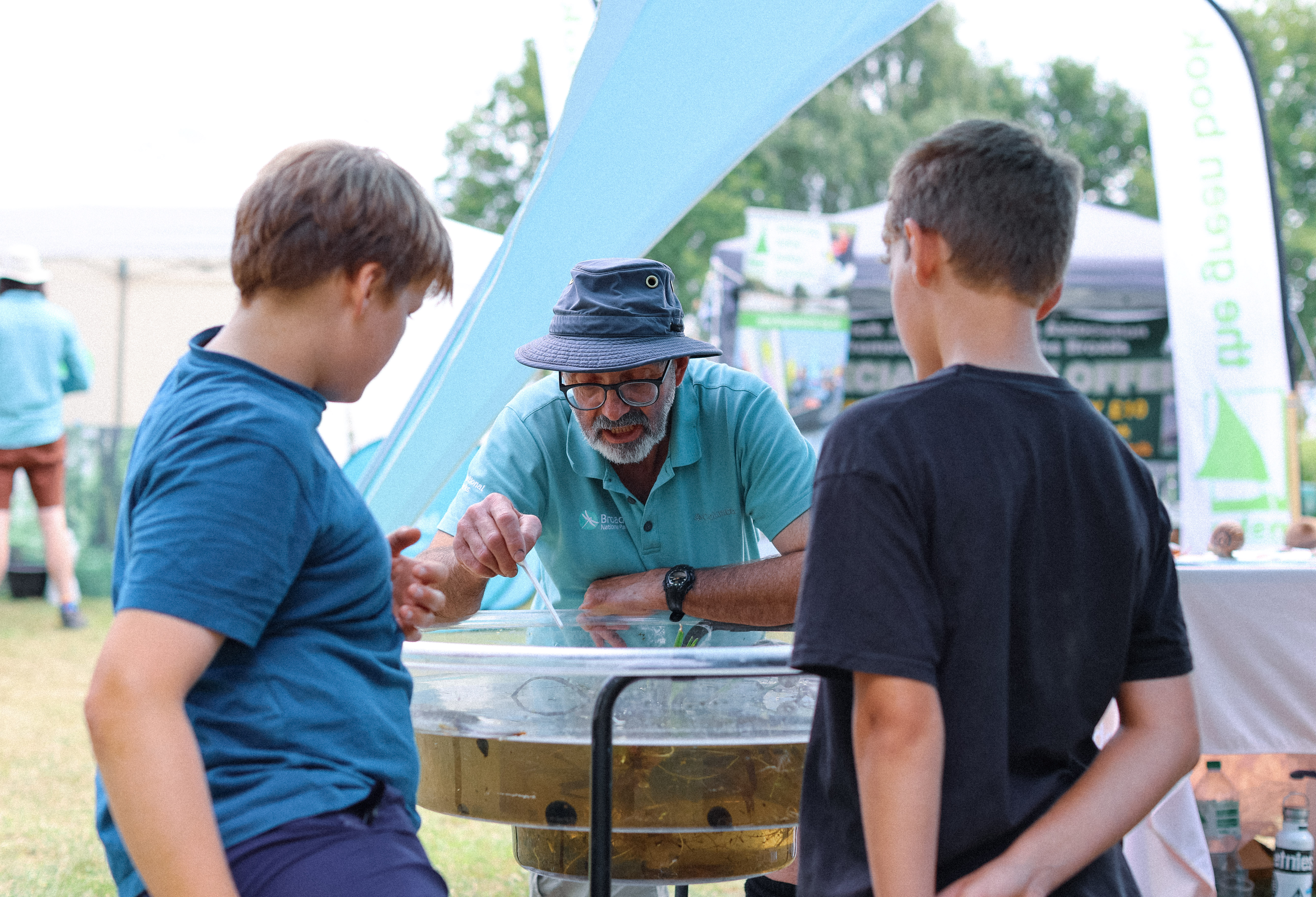 An outdoor nature activity where a guide shows two children aquatic wildlife in a clear water tray under a marquee, with event tents and banners in the background.