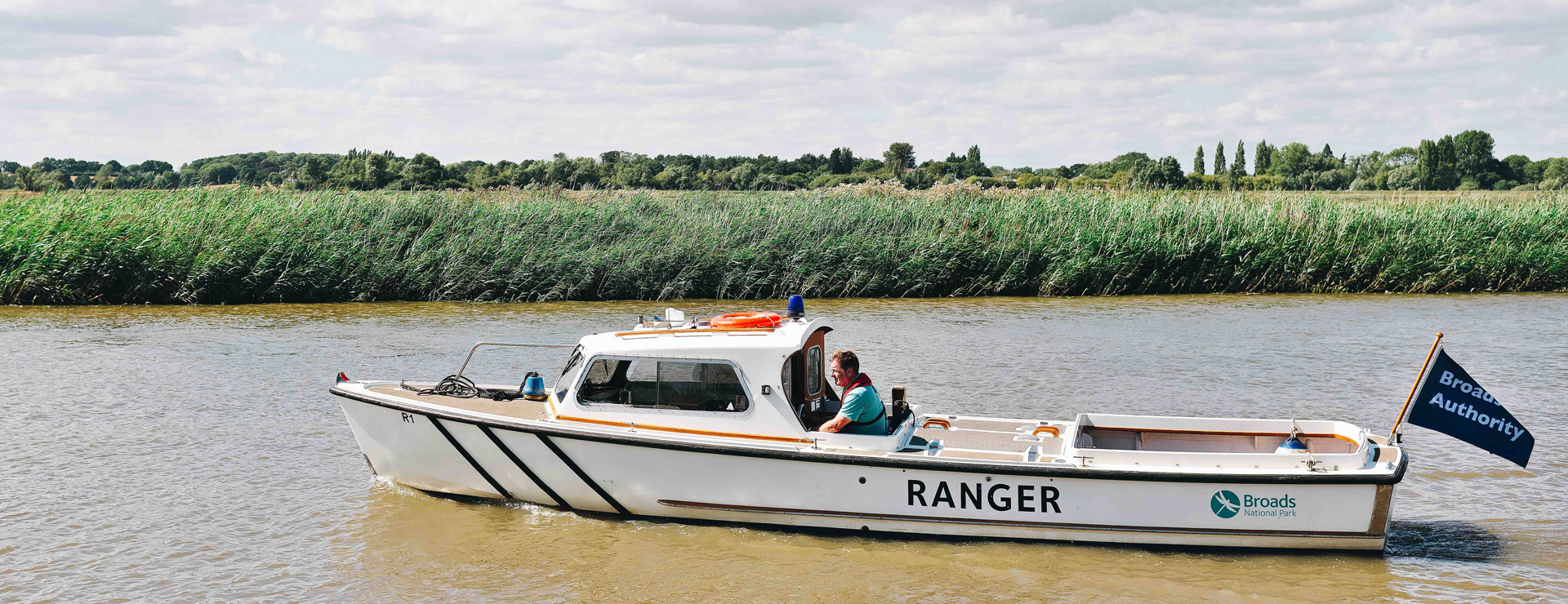 A Broads Authority Ranger in a white patrol boat with the word RANGER on the side, along with the Broads Authority teal dragonfly logo, travels down the river from right to left. The boat is being helmed by a man wearing a teal polo shirt and life jacket. In the background the rest of the river, along with the green fringes of reeds/sedge on the edge of the water. Trees and clouds can be seen in the distance.