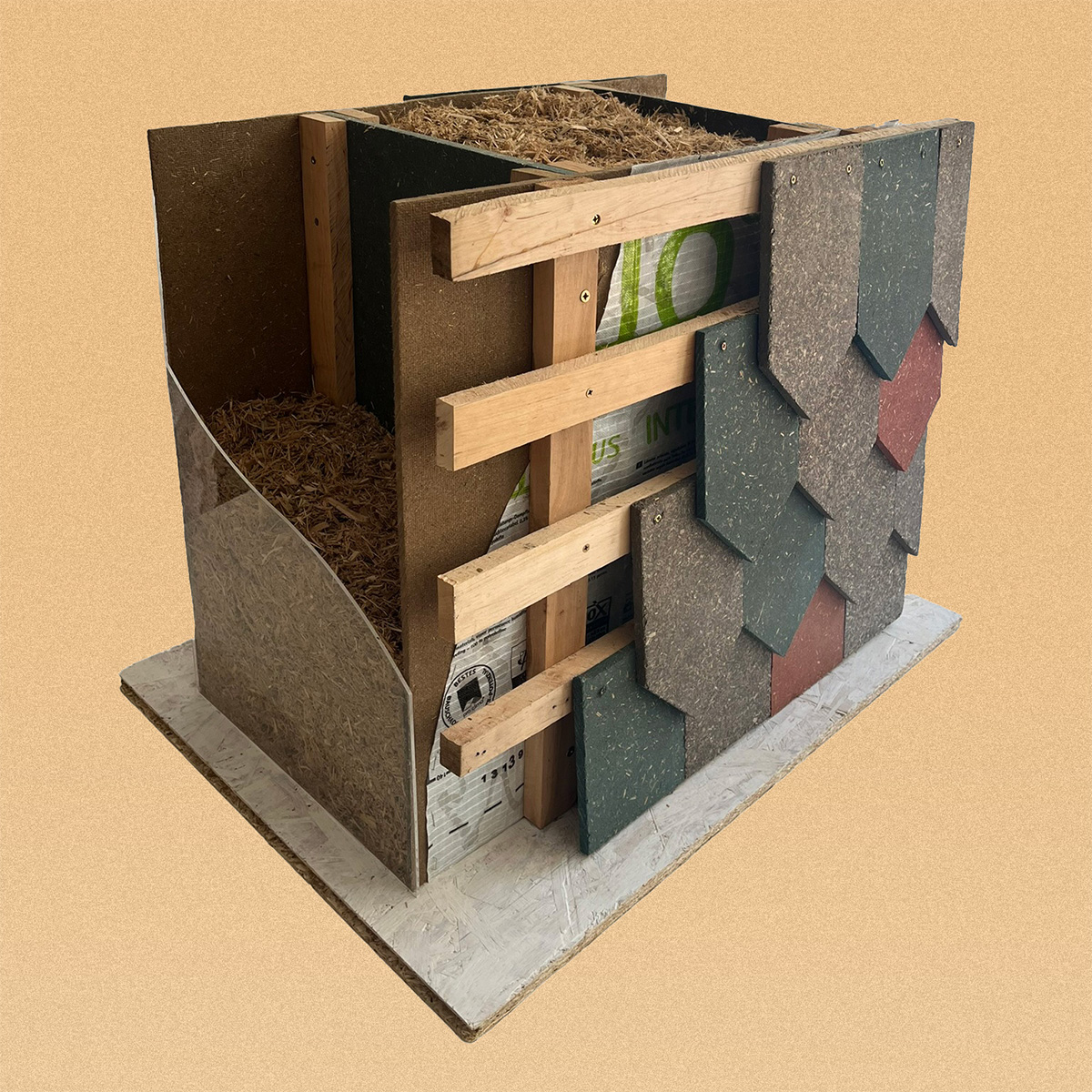 Demonstration wall build by Hudsons Architects A small architectural demonstration model showing a cross-section of a wall assembly, with wooden framing, insulation made of shredded plant material, and exterior cladding pieces in different colours arranged like shingles.