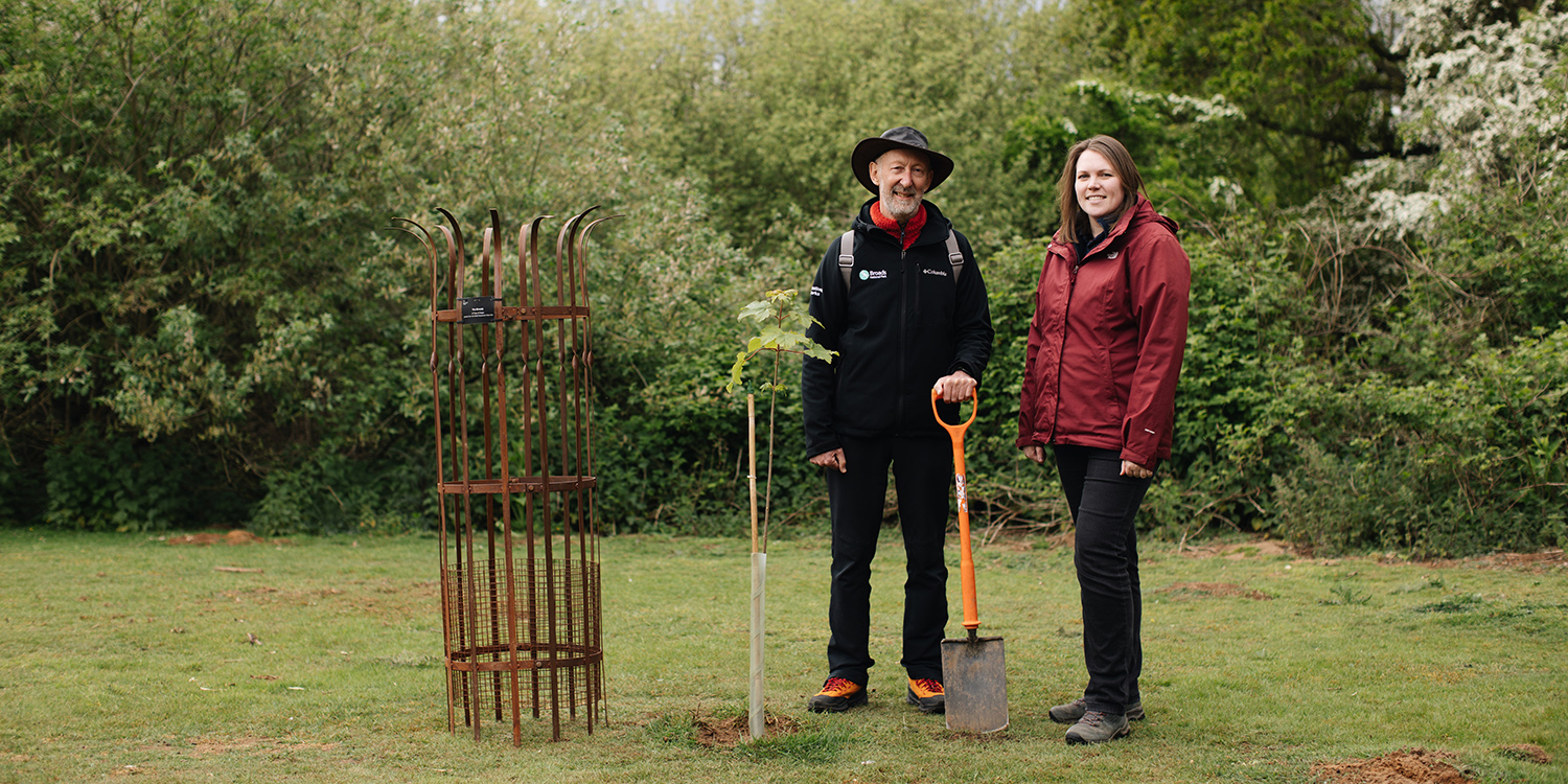 Two people stand in a green outdoor setting beside a newly planted young tree and a metal tree guard; one holds a spade, with dense trees and bushes behind them.&rdquo;