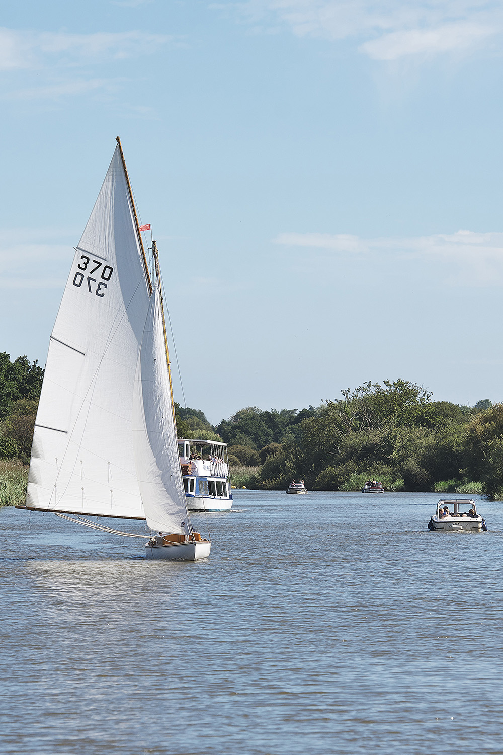 Traditional white-sailed yacht sailing along a broad river on a sunny day. Several motorboats share the waterway, while trees and reeds line the banks beneath a clear blue sky.