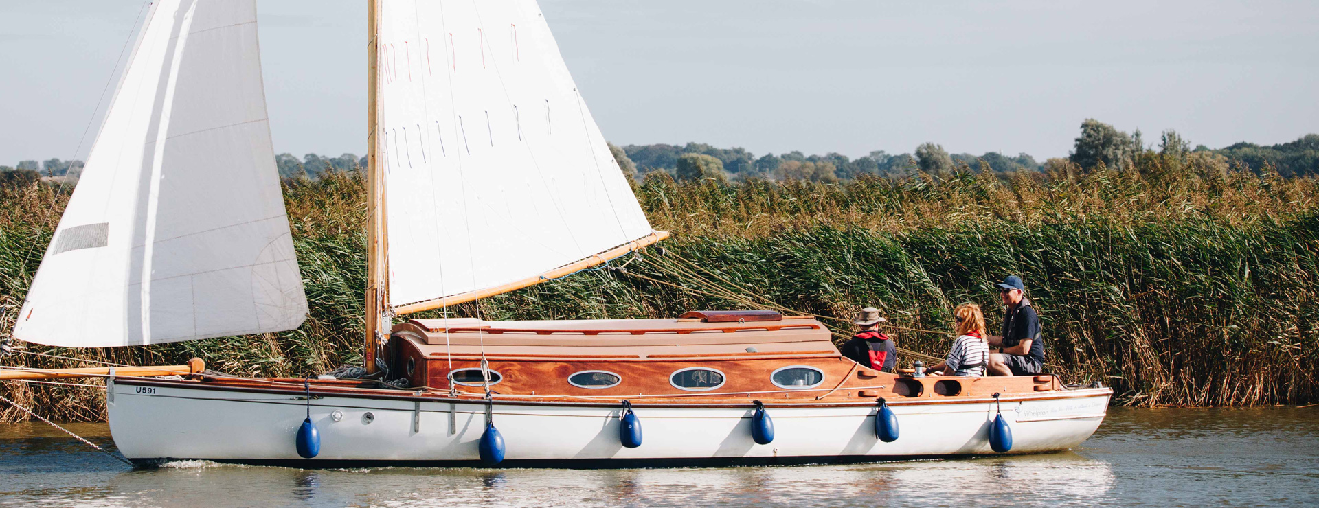 A wooden sailing boat with a large white sail travels down the river on a sunny day, with reeds visible in the background. The boat is being crewed by two men and a woman who are all wearing life jackets. The boat is travelling from right to left across the frame. Hazy clouds can be seen in the distance.