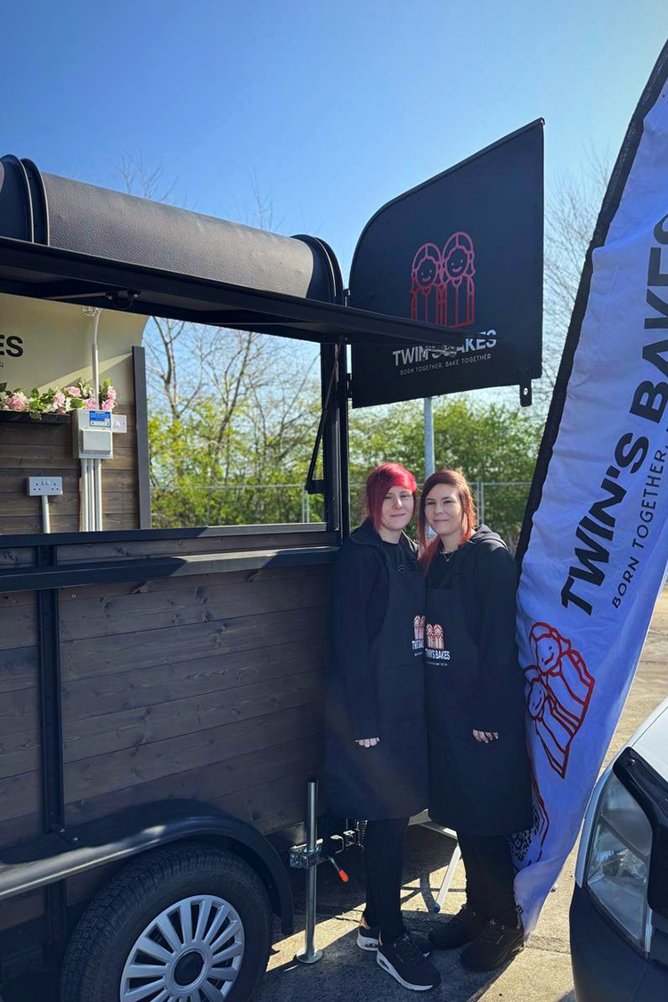 Twin's Bakes will be opening up at Acle soon Two members of Twin Bakes standing beside their mobile bakery trailer at Acle, with branded signs and flag visible.
