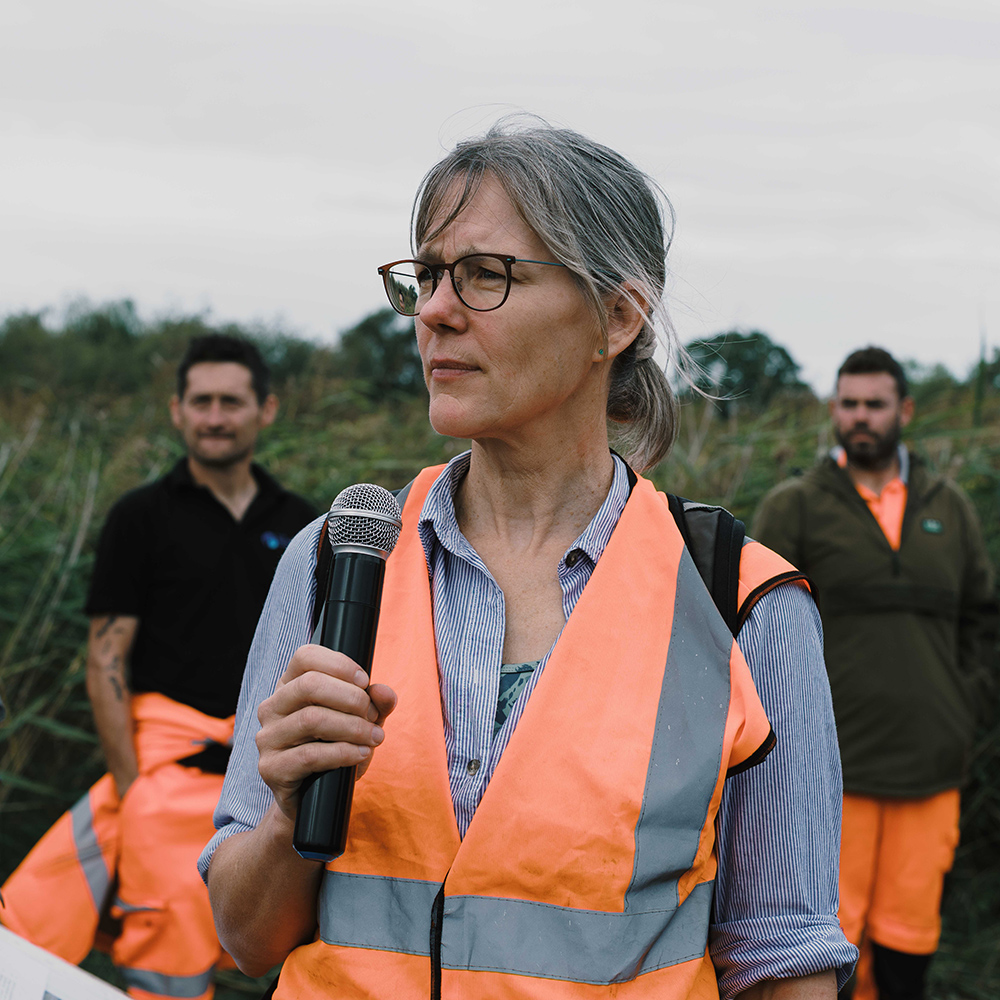 Environmental Policy Adviser Andrea Kelly speaks at the event Andrea Kelly, wearing a high-visibility vest, holds a microphone and speaks at a wildfire awareness event, with two participants standing behind her.