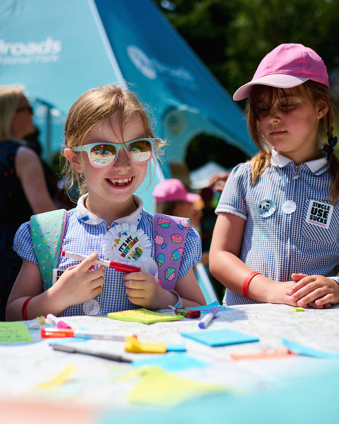 Two young girls in blue checkered school uniforms and colorful backpacks participate in a craft activity at an outdoor event. One girl, wearing sunglasses and a birthday badge, smiles while holding a red pen. The other, in a pink cap, looks on. A table in front of them is covered with bright paper, pens, and markers. A “Broads National Park” tent is visible in the background.