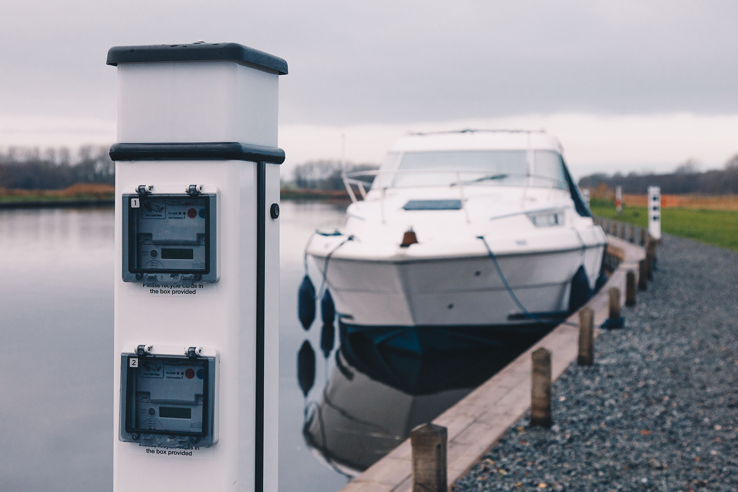 An electric charging point at a Broads Authority mooring A close-up image of a metal electric charging point on the Broads, with a motor cruiser visible in the distance