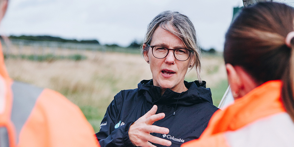 Andrea Kelly, Broads Environment Policy Adviser A woman wearing glasses and a black Columbia jacket speaks to two people in orange safety jackets in an outdoor setting, possibly a field or work site.