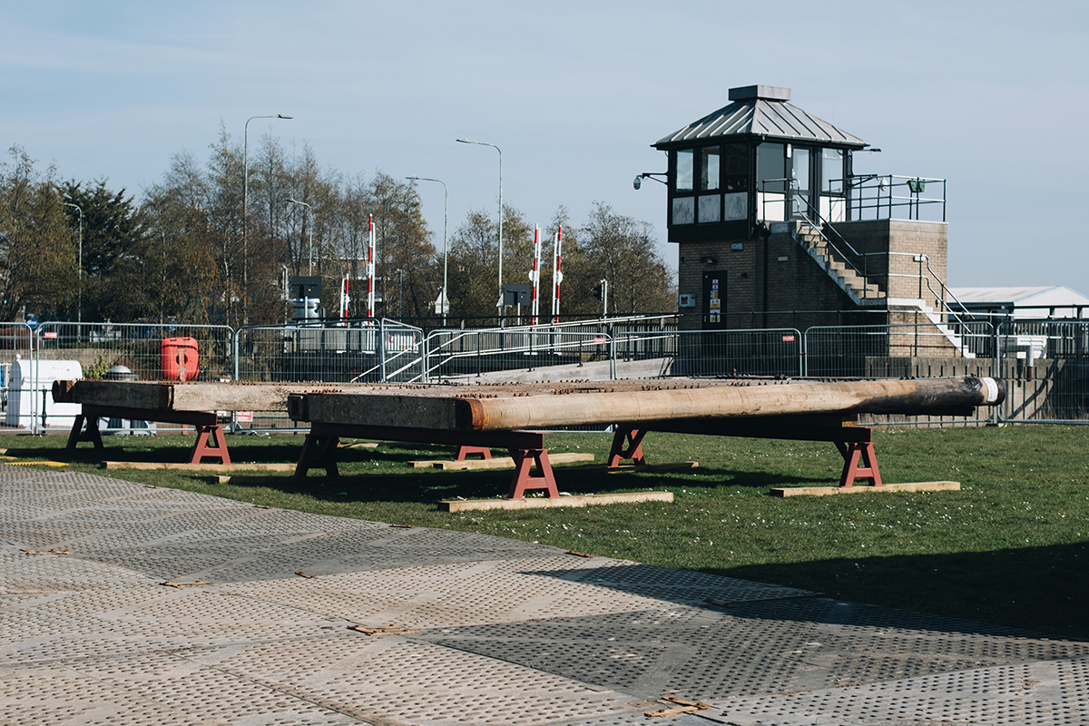 A close-up view of removed wooden lock gates placed on metal supports during repair works at Mutford Lock. The gates appear weathered and worn, with visible bolts and metal reinforcements. In the background, there is a control building with a metal staircase, traffic barriers, and fencing around the construction site.