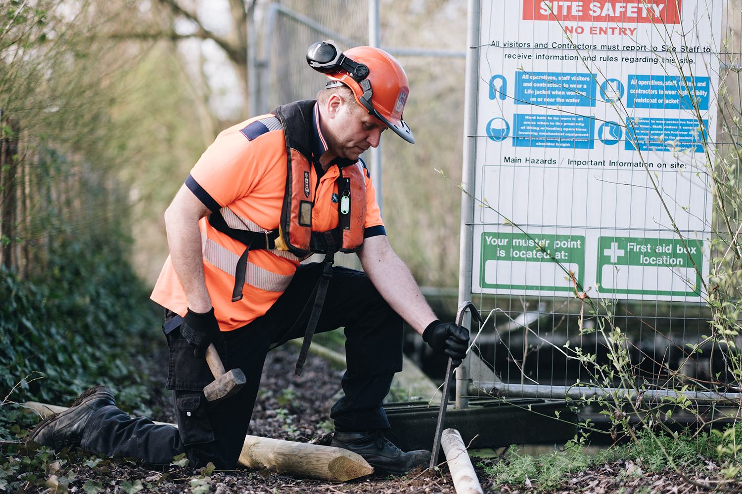 Working at Hoveton Riverside Park A Broads Authority worker wearing orange PPE uses a hammer to hit something into the ground next to some metal HERAS fencing