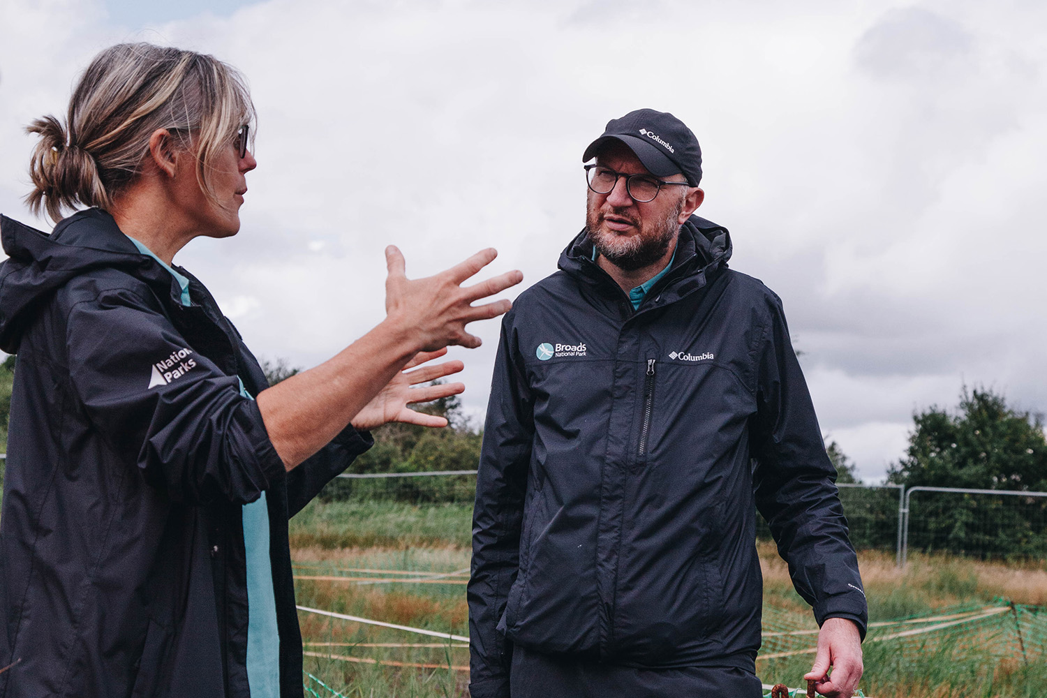Two members of the Broads peatland team at the Horsey wetland site Two Broads Authority staff members wearing outdoor jackets stand in a grassy field having a conversation. The person on the left gestures with both hands while speaking, and the person on the right listens with hands in pockets, wearing glasses and a cap. A cloudy sky and some fencing are visible in the background.