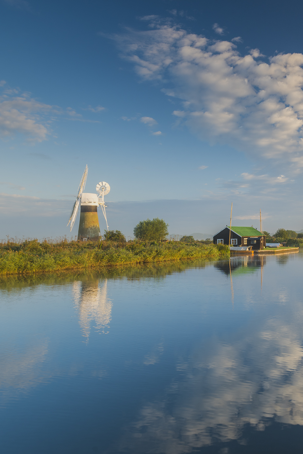 The Broads on the River Thurne, by Tom Mackie A scenic view of the Broads at Thurne, with a traditional windmill and a small riverside cottage reflected in the calm water under a bright blue sky with scattered clouds.