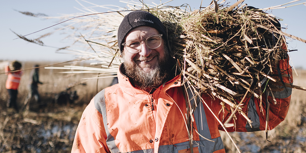 A man wearing an orange high-vis jacket holding a bundle of reeds.