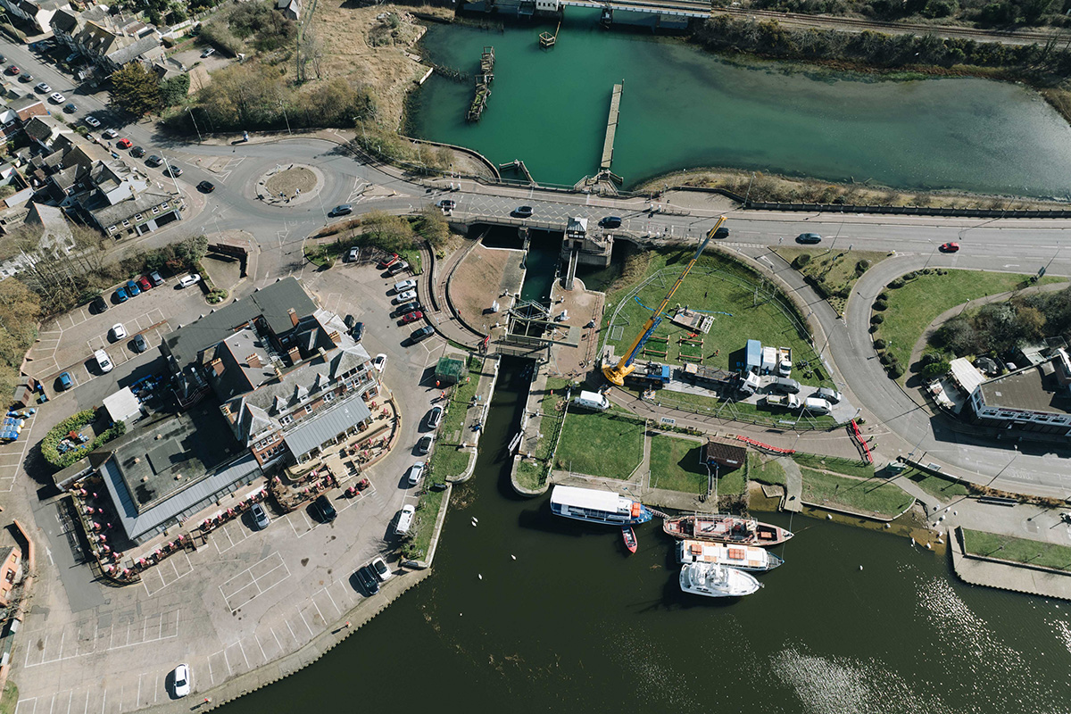 Aerial view of Mutford Lock showing ongoing repair works. Construction equipment and materials are visible near the lock, with workers actively involved in the maintenance. The lock connects Oulton Broad to Lake Lothing, surrounded by moored boats and a marina in the background. Nearby buildings, a parking area, and a busy road with cars are also visible.