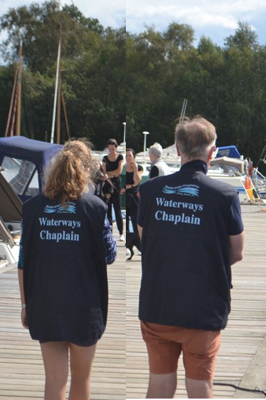 Waterways Chaplains wear a distinctive gilet Two people wearing navy blue vests with 'Waterways Chaplain' printed on the back walk along a marina dock, with boats moored on either side and trees in the background.