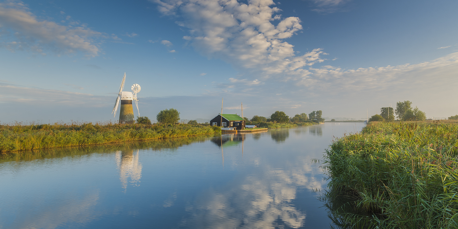 Scenic view of the Broads at Thurne, with a traditional windmill and a small riverside cottage reflected in the calm water under a bright blue sky with scattered clouds.