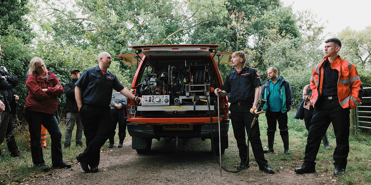 Participants at the wild fire awareness event in the Broads.jpg A group of people stand around the open rear of a red emergency response vehicle parked on a woodland track. Two crew members demonstrate equipment stored in the vehicle, including hoses, pumps and control panels, while onlookers gather nearby among trees and vegetation.