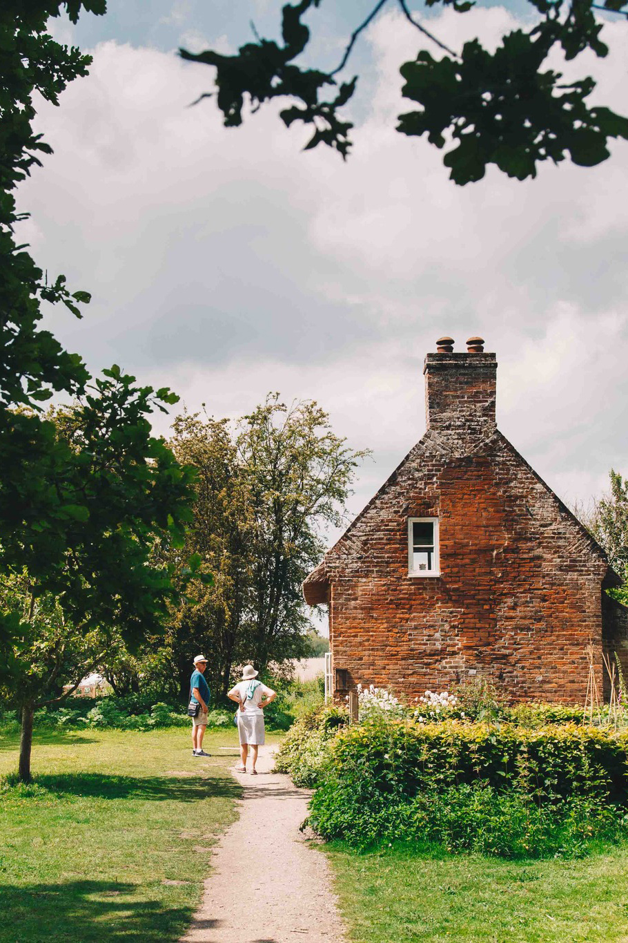 Two people standing on a garden path in front of a quaint red-brick cottage with a single white-framed window and a tall chimney. The cottage is surrounded by lush greenery, flowering bushes, and trees under a partly cloudy sky.