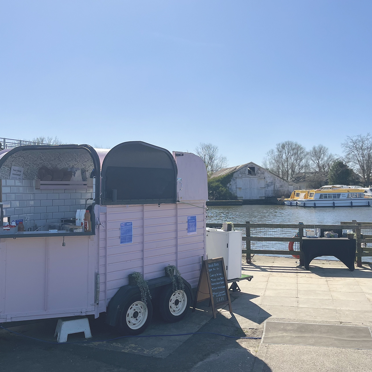 A pastel pink food trailer with a serving window open, set up by a riverside on a sunny day. The trailer has white tiled walls inside and a small chalkboard menu outside. In the background, there's a calm river with moored yellow and white boats, an old weathered building, and leafless trees under a clear blue sky.