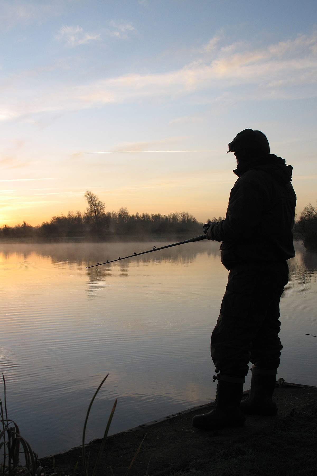 A silhouette of a person fishing by a calm lake at sunrise or sunset, with mist hovering over the water and soft pastel tones in the sky.
