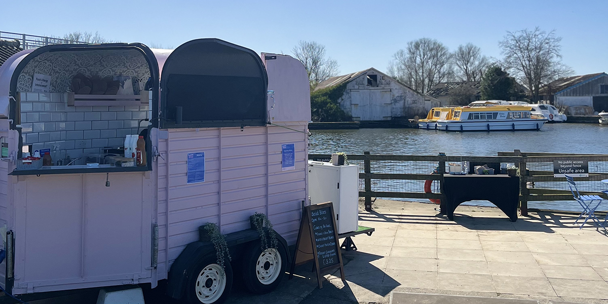 A pastel pink food trailer with a serving window open, set up by a riverside on a sunny day. The trailer has white tiled walls inside and a small chalkboard menu outside. In the background, there's a calm river with moored yellow and white boats, an old weathered building, and leafless trees under a clear blue sky.