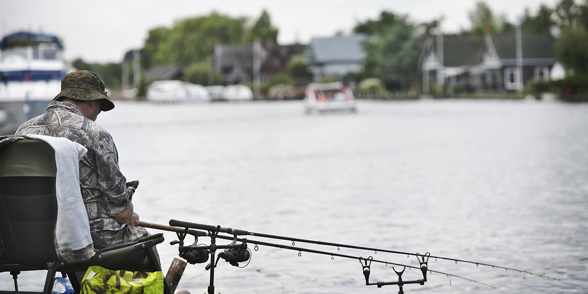 An Angler seated by a river with fishing rods extended into the water, wearing camouflage clothing and a bucket hat, with boats and houses in the background.