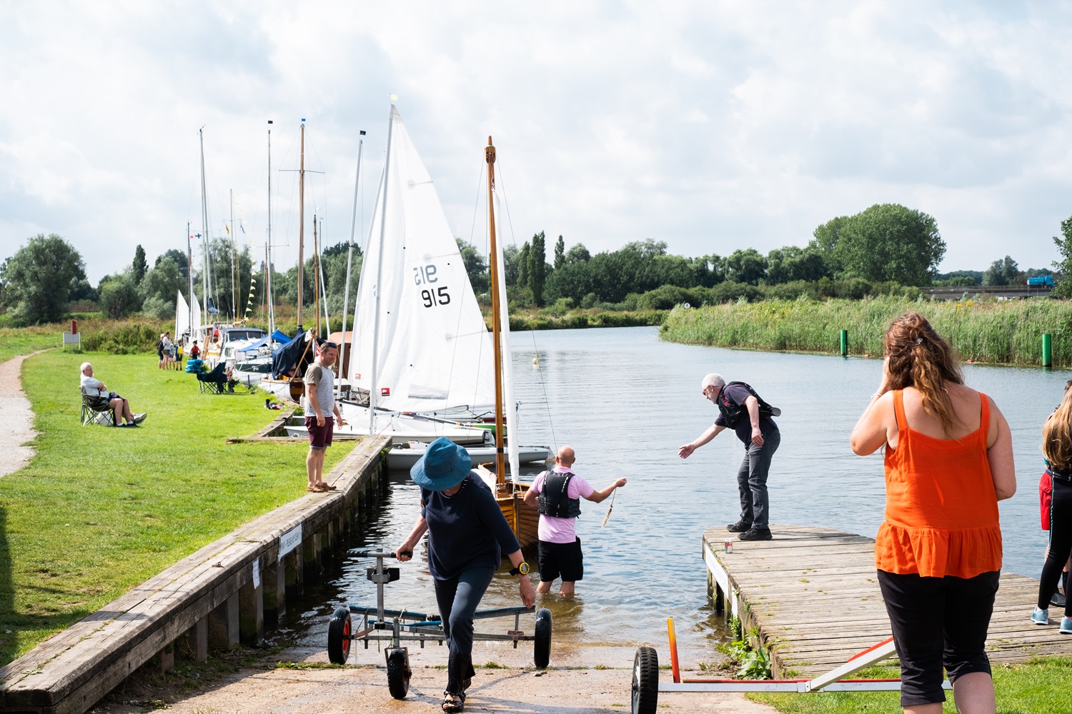 People launching and retrieving small sailboats at a riverside slipway on a sunny day. A woman in a blue hat pulls a boat trolley up the ramp, while others assist a boat in the water. Several sailboats are moored along the grassy bank, and more people relax or help nearby. Lush green trees and reeds line the opposite riverbank under a partly cloudy sky.