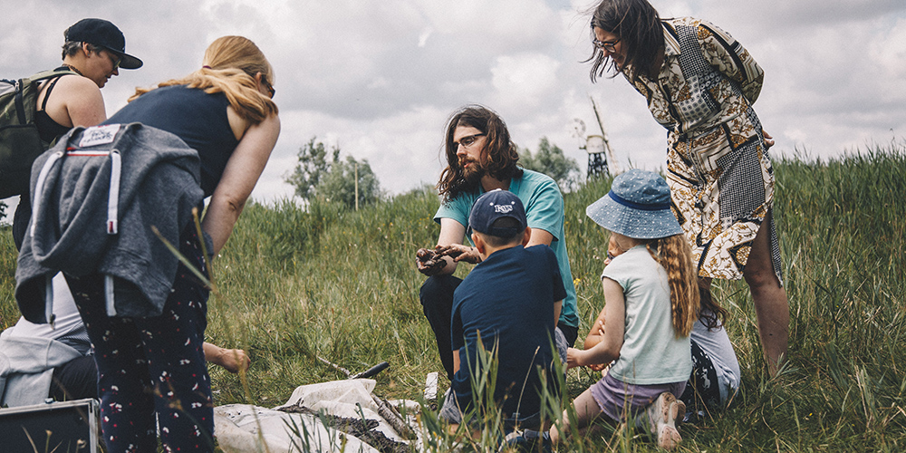 A group of people, including adults and children, are gathered outdoors in a grassy area on a cloudy day. A man with long hair and a beard sits in the center showing something in his hands, while others watch attentively. Some people are standing, and others are kneeling or sitting on the grass, suggesting an educational or nature activity.
