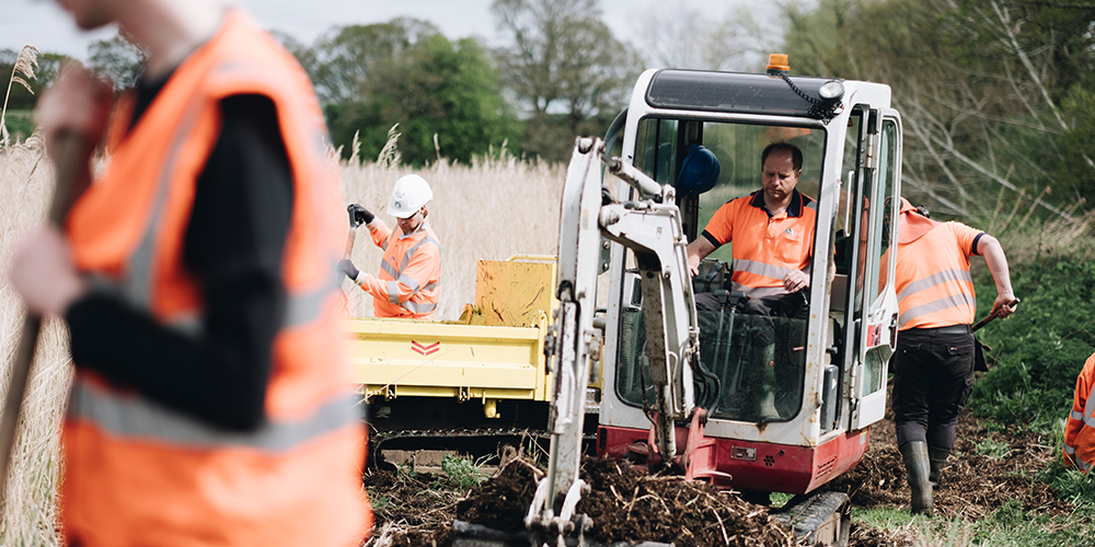 Workers wearing high-visibility orange vests and safety gear operate machinery and dig in a marshy outdoor area, carrying out land management or construction work.