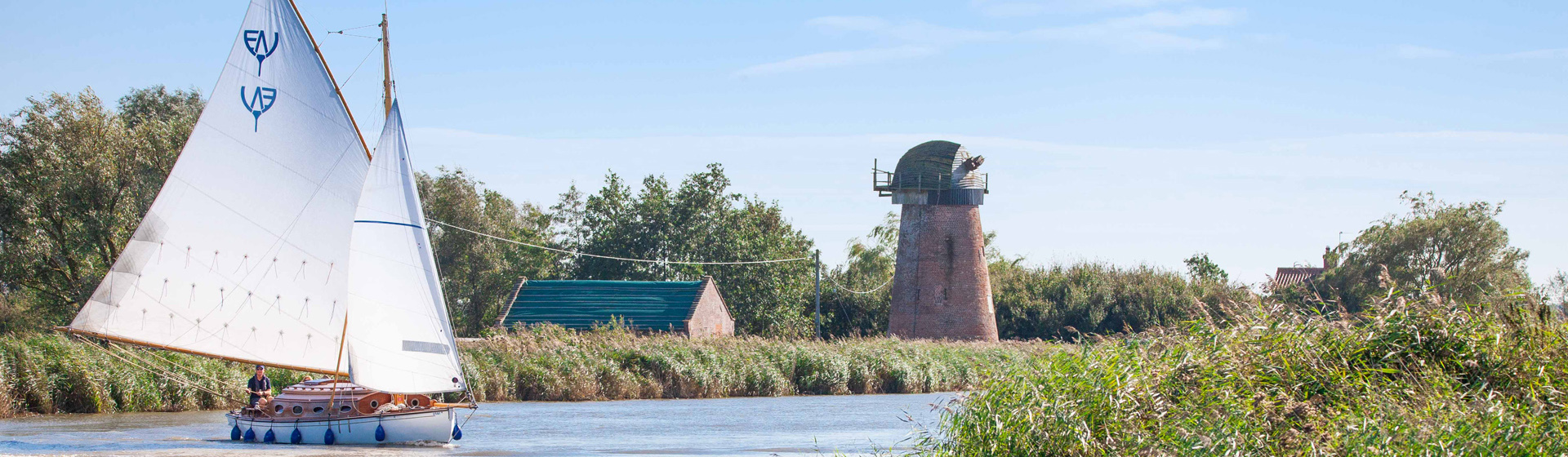 A small sailboat with white sails glides along a calm river bordered by tall reeds. In the background, a rustic brick mill with a weathered cap stands among trees under a clear blue sky.