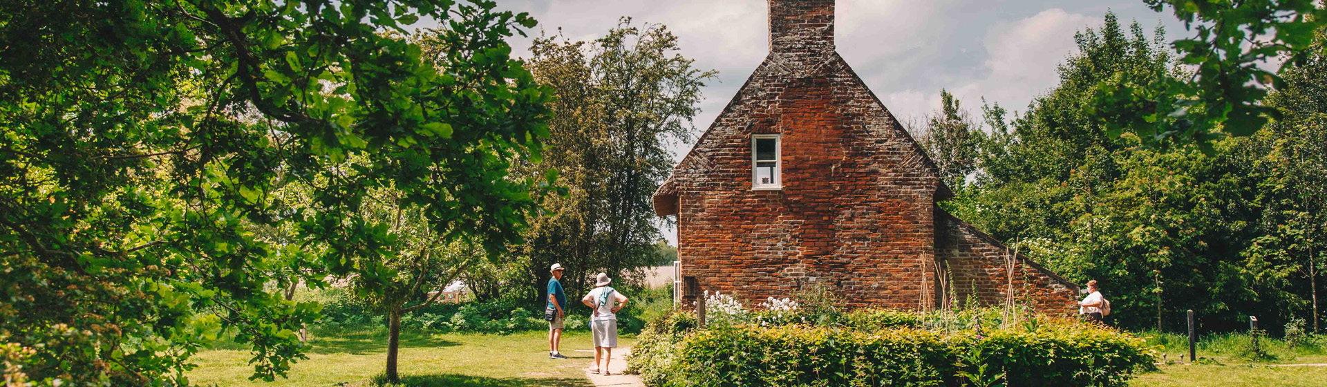 A small historic brick cottage with a tall central chimney at How Hill, surrounded by lush green trees and gardens. A few visitors walk along the grassy path in front of the cottage on a bright, partly cloudy day.